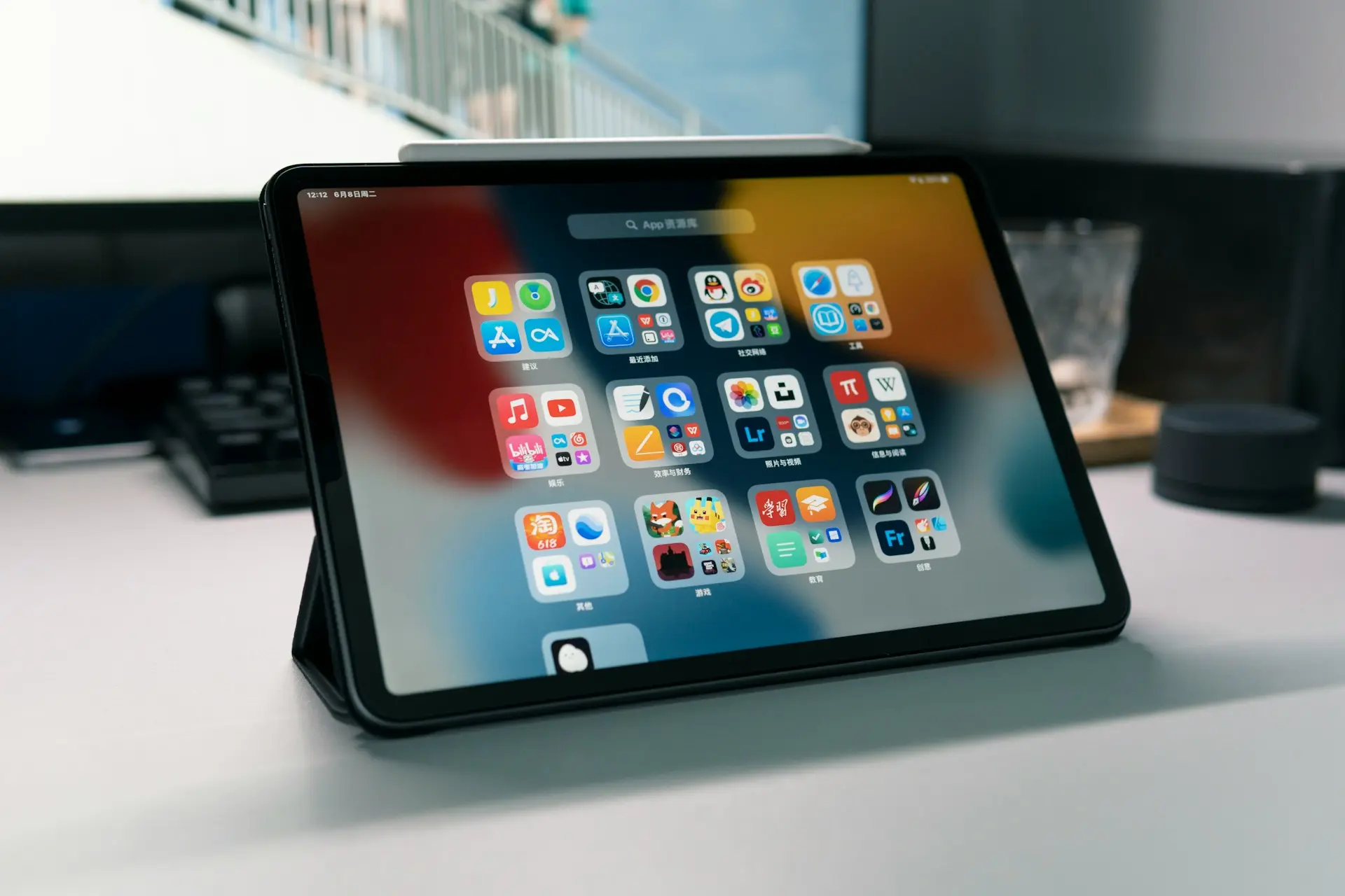Tablet iPad apoyada en su funda sobre un escritorio de trabajo, mostrando la pantalla de inicio con carpetas de aplicaciones organizadas y un lápiz digital acoplado en la parte superior.